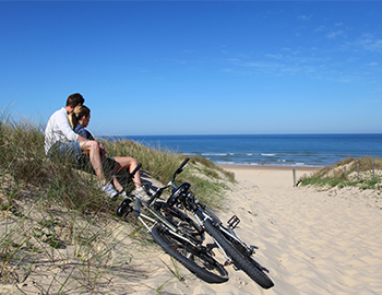 Paar sitzt in den Dünen und schaut über Strand und Meer, daneben zwei Fahrräder