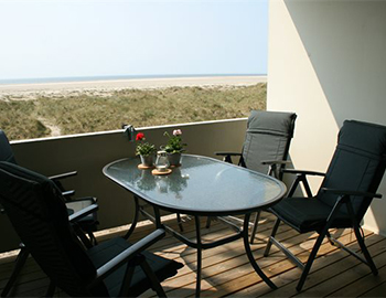 Balkon mit Tisch und vier Stühlen. Vom Balkon aus gibt es Aussicht über die Dünen, den Strand und das Meer